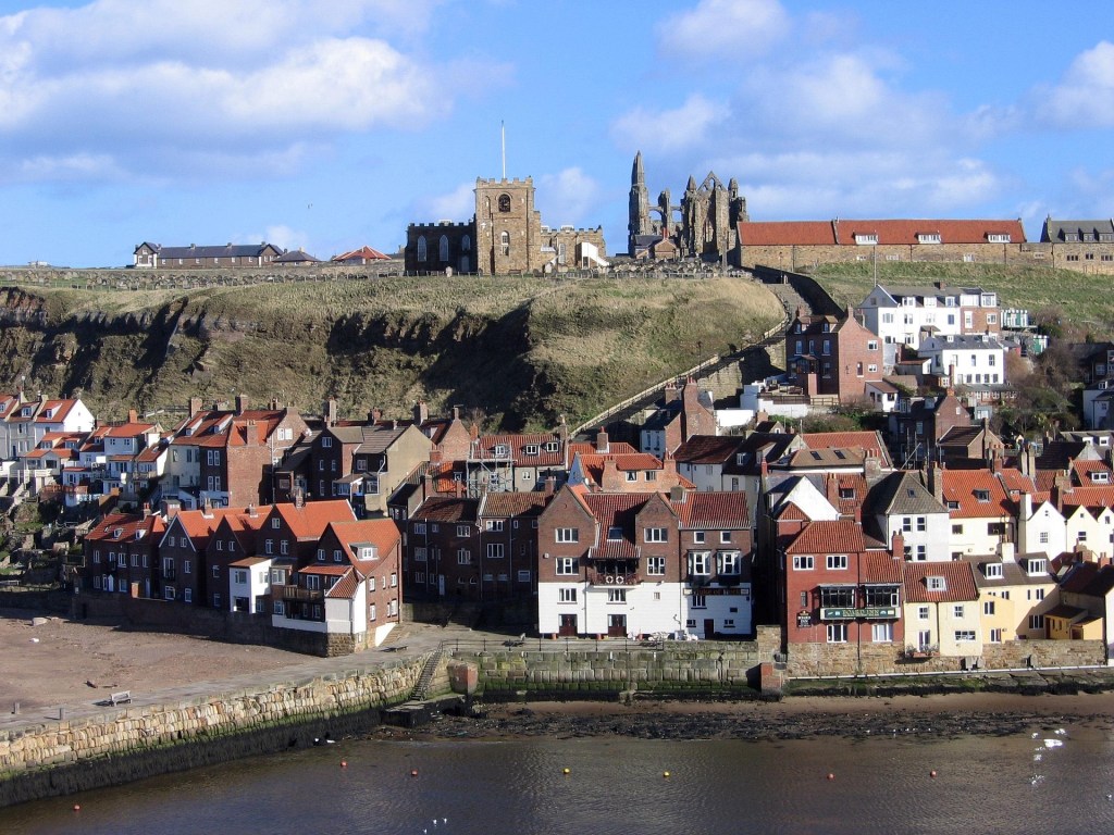 A daytime exterior photograph of Whitby's East Cliff, showing the historic red-roofed town buildings along the harbour wall, with St Mary's Church and the dramatic ruins of Whitby Abbey standing on the headland above under a blue sky with light clouds.